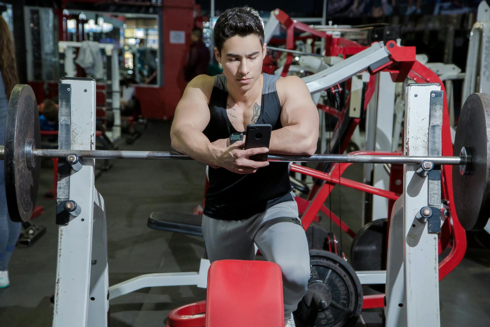 A young man resting in a gym while using a mobile phone beside weightlifting equipment.